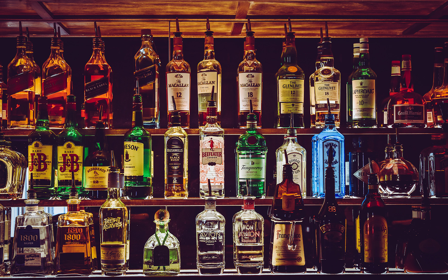 Dark backdrop with variety of brands of alcohol bottles lit up in rows.
