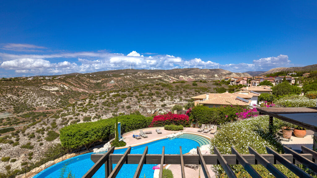 Zephyros Village communal pool on Aphrodite Hills Resort, looking from the pool water toward the sunbathing terrace with mountain view in distance. Presented by Aphroditerentals.com