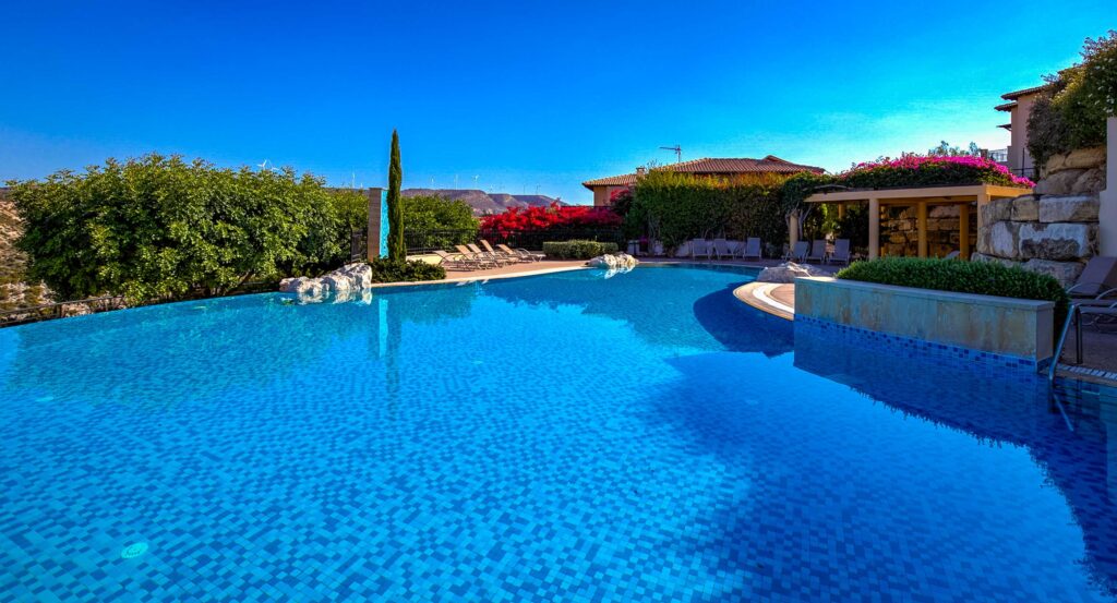 Zephyros Village communal pool on Aphrodite Hills Resort, looking from the pool water toward the sunbathing terrace with mountain view in distance. Presented by Aphroditerentals.com