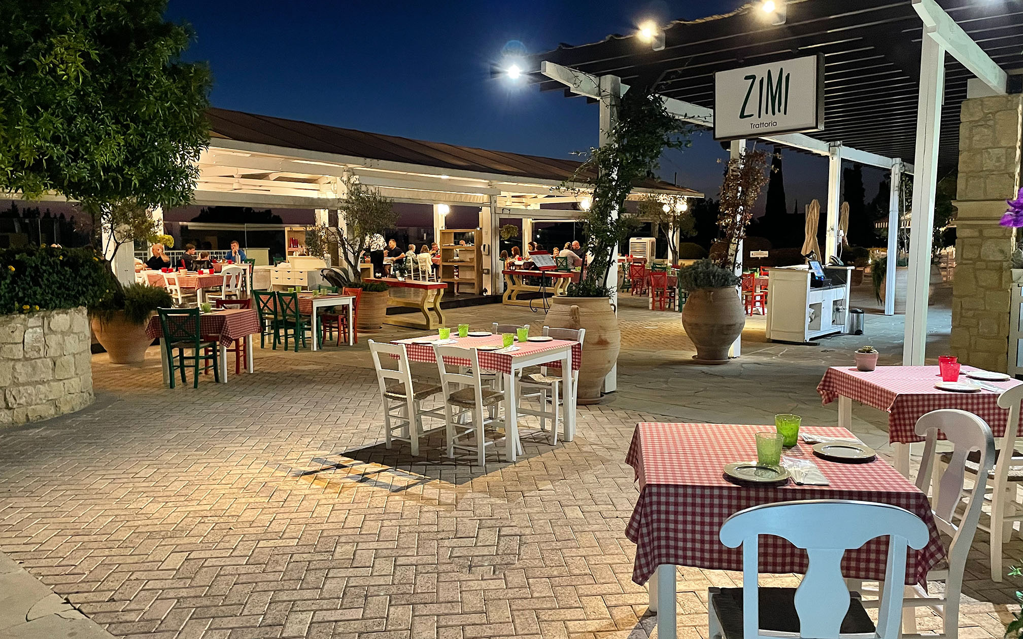 View of the Zimi Italian restaurant on Aphrodite Hills Resort, tables and chairs set up, lit up in dark evening.