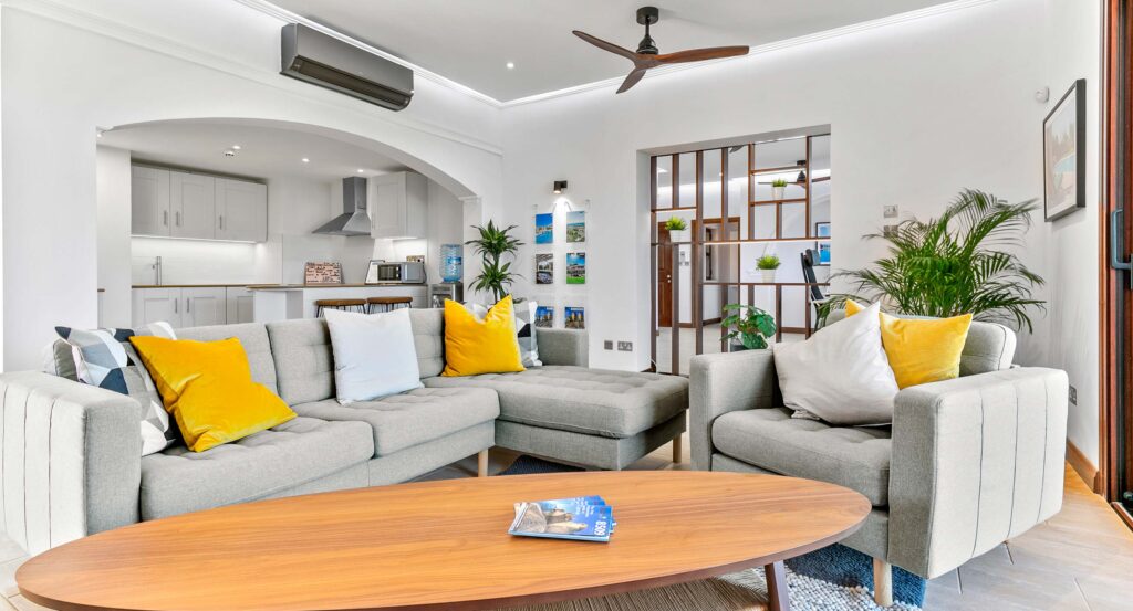 View inside the departure lounge living area and kitchen of Aphroditerentals.com, showing comfortable grey sofas, wooden table kitchen area, with wooden ceiling fan. Aphrodite Hills Resort, Cyprus.