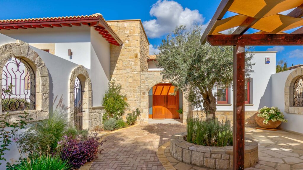 Courtyard entrance of Villa Agapi - stone walls and window frames, olive tree, pergoda with shade and large wooden front door.