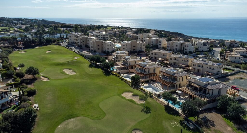 Aerial view of Dionysus Village on Aphrodite Hills - showing the golf course, surrounded by cream properties, and some swimming poosl, with the sea in the background.