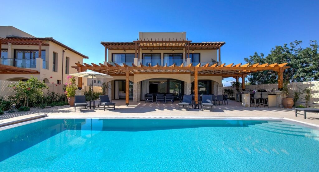 Photograph of a large private pool infront of a large private villa, with seating, parasol, pretty plants on terrace, and blue skies behind.