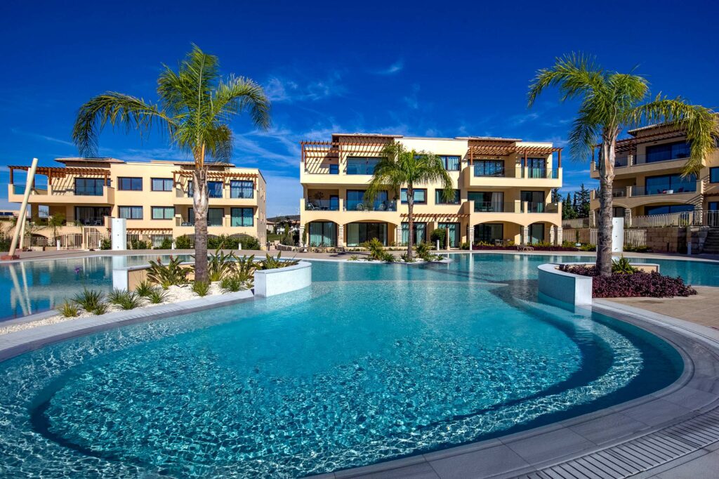 Photograph of Large communal pool with palm trees, and apartment blocks in the background. Big blue sky in the background.