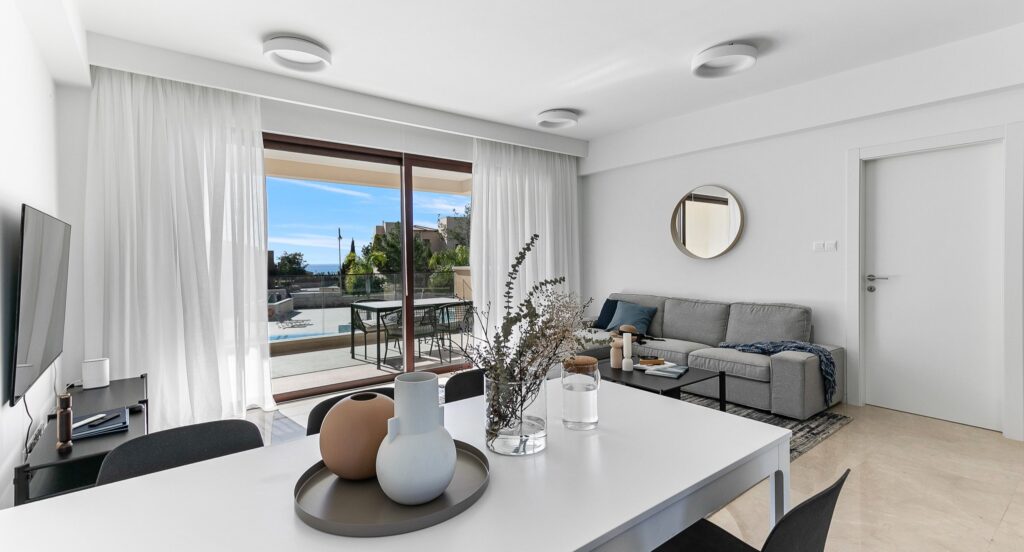 Photograph of an apartment living area, with dining table and chairs in the foreground, sofa and TV behind, large patio doors opening out to a balcony and views of the pool and Mediterranean Sea.